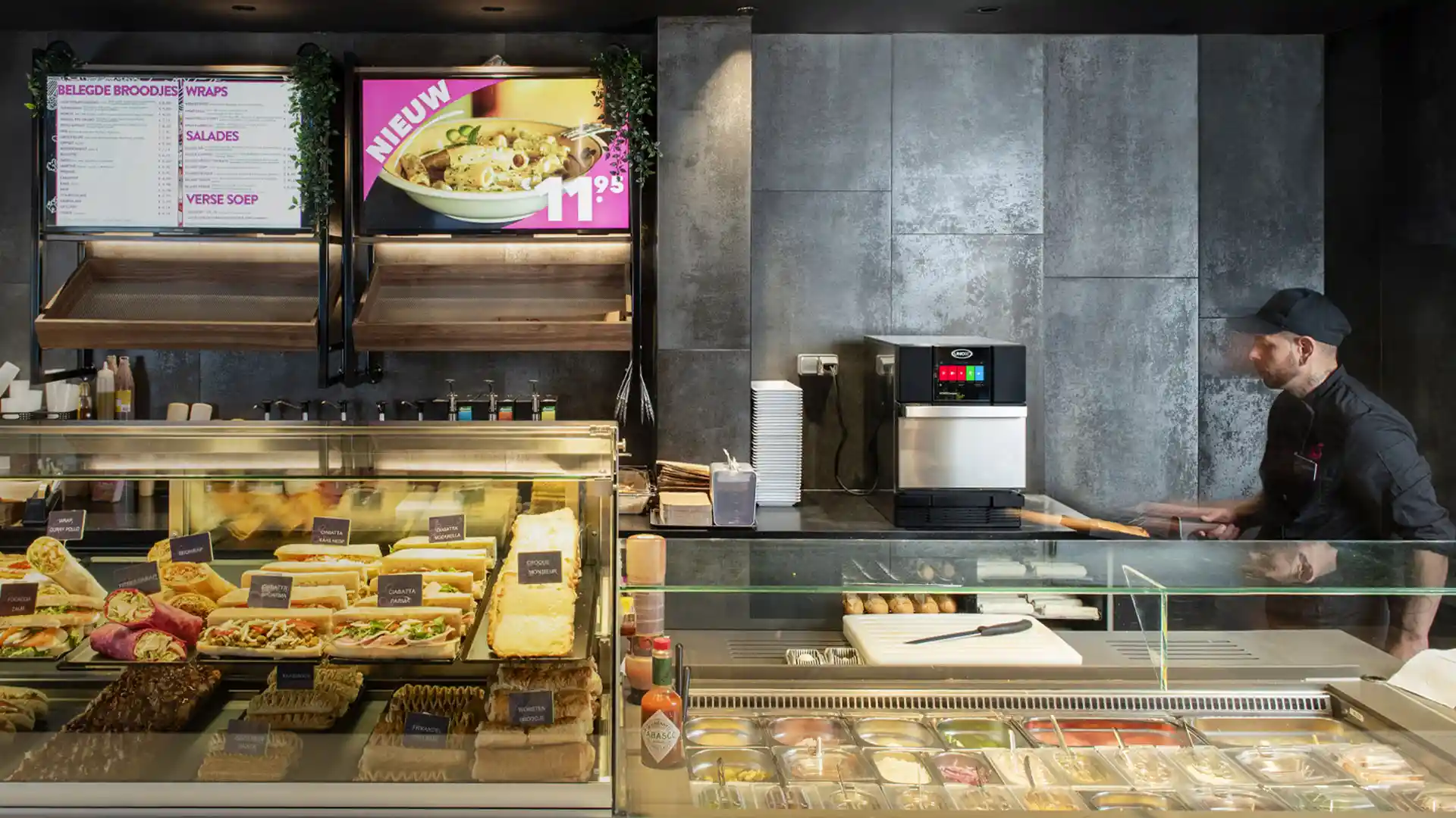 Funcionário de restaurante de serviço rápido (QSR) olhando para um forno de aceleração Unox atrás do balcão com wraps, sanduíches e saladas.