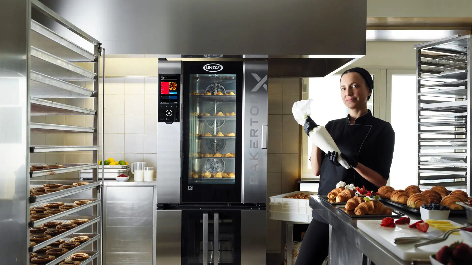 Um confeiteiro na cozinha, ao lado de um forno combinado Unox com doces no interior, segurando um saco de confeitar. Os doces estão expostos em um balcão próximo.