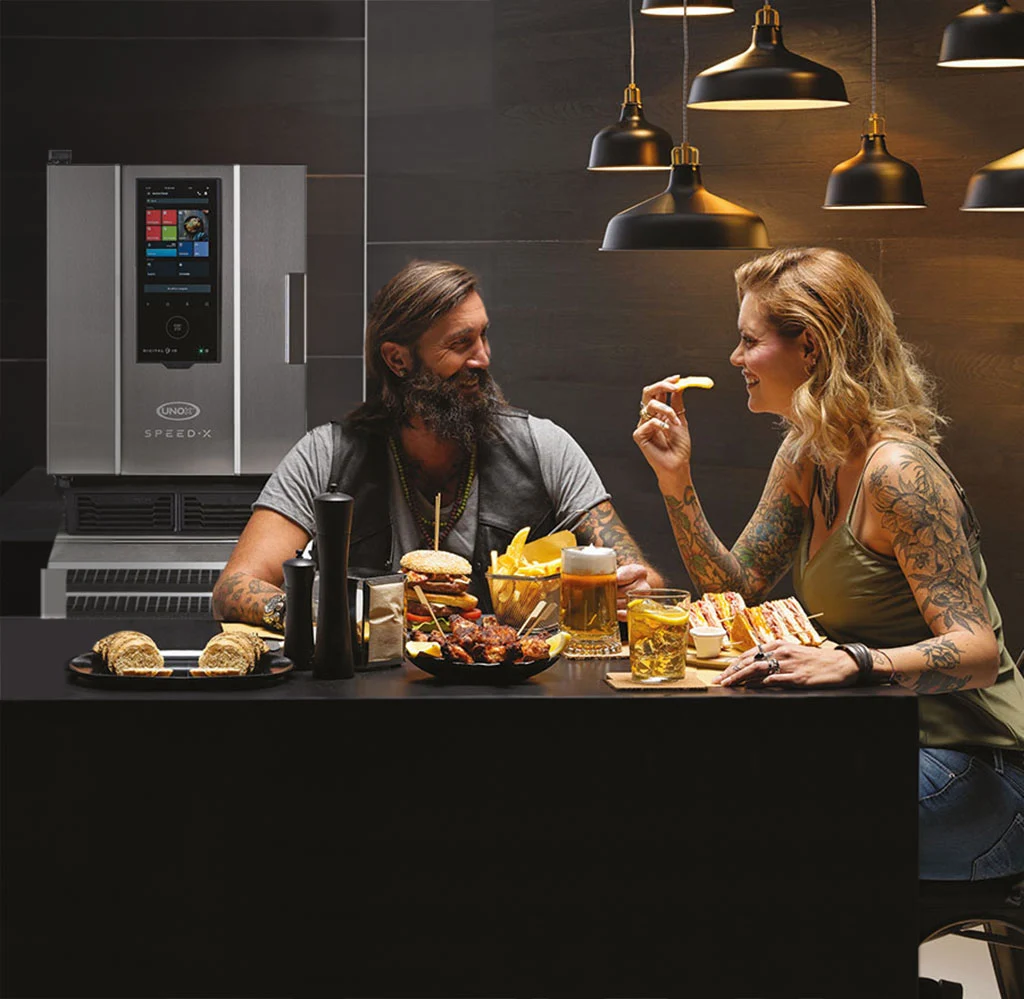 Um homem e uma mulher sorridentes, ambos com tatuagens, estão sentados à mesa com hambúrgueres, batatas fritas, sanduíche e outros alimentos, cerveja, um coquetel e um forno de aceleração Unox atrás deles.