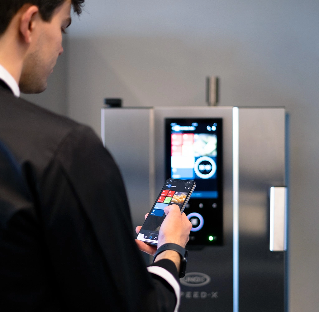 Homem com dólmã de chef preta segurando um smartphone e pressionando alguns botões, voltado para um forno rápido Speed-X Unox, controlando o forno pelo telefone.