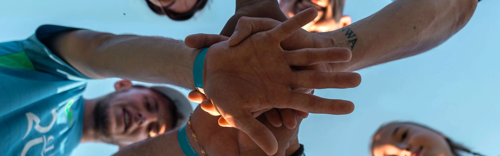 Um grupo de pessoas olhando para baixo em direção à câmera, colocando as mãos umas sobre as outras, simbolizando trabalho em equipe.